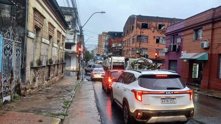 Camiones de recolección de basura en una calle lluviosa, con vehículos en fila y edificios de fondo, en un día nublado.