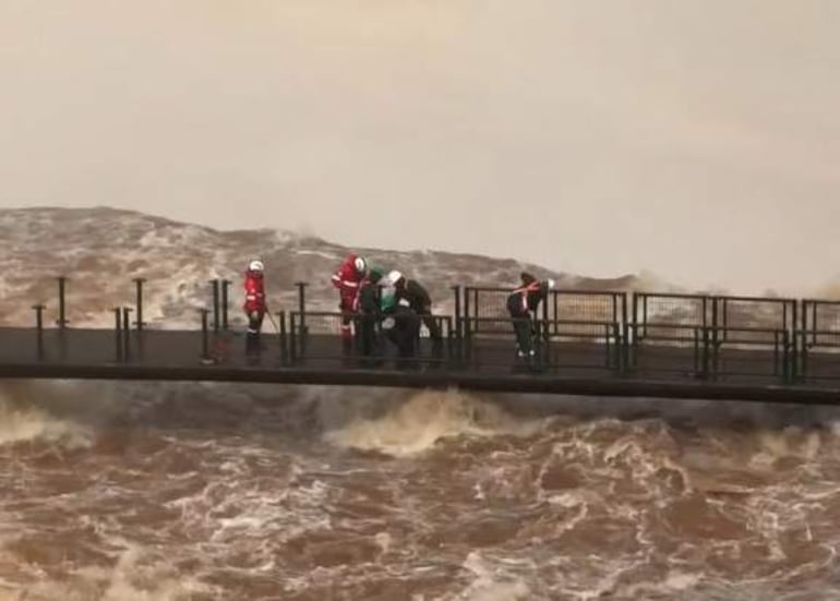 Cuatro personas con trajes de rescate en plataforma, ayudando a un individuo en medio de aguas turbulentas bajo un cielo nublado.