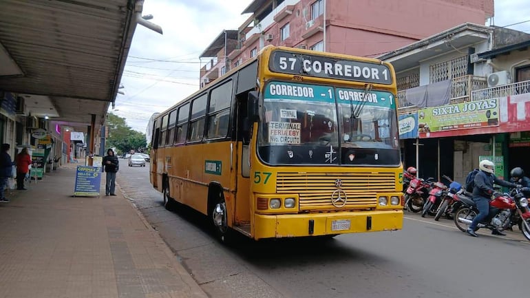 Autobús amarillo con la inscripción '57 CORREDOR 1', personas en la vereda mirando teléfonos en un ambiente urbano.