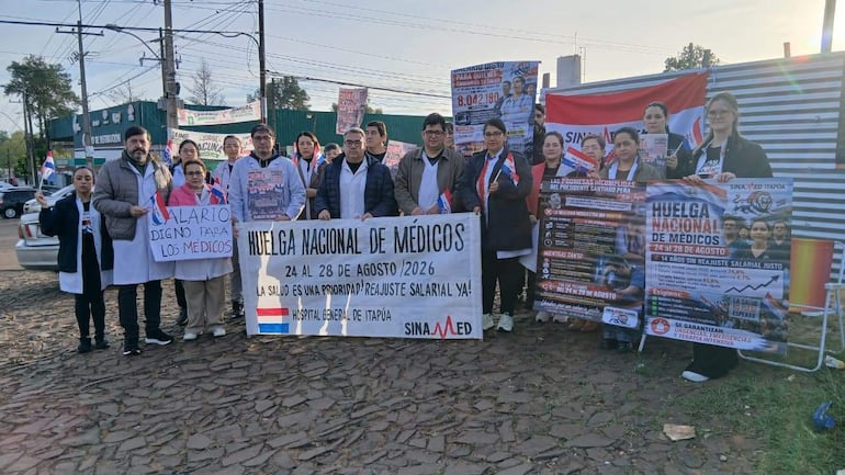 Grupo de médicos en batas blancas sostiene pancartas sobre huelga nacional, en un ambiente urbano con edificios y vehículos de fondo.