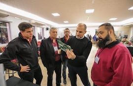 Cuatro hombres en sala de conferencia, vestidos formalmente, discuten sobre un dispositivo técnico perteneciente a la máquina de votación; uno sostiene una placa y otros observan atentos.