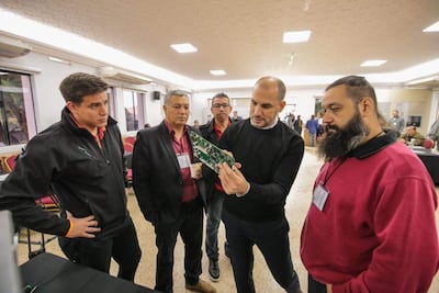 Cuatro hombres en sala de conferencia, vestidos formalmente, discuten sobre un dispositivo técnico; uno sostiene una placa y otros observan atentos.