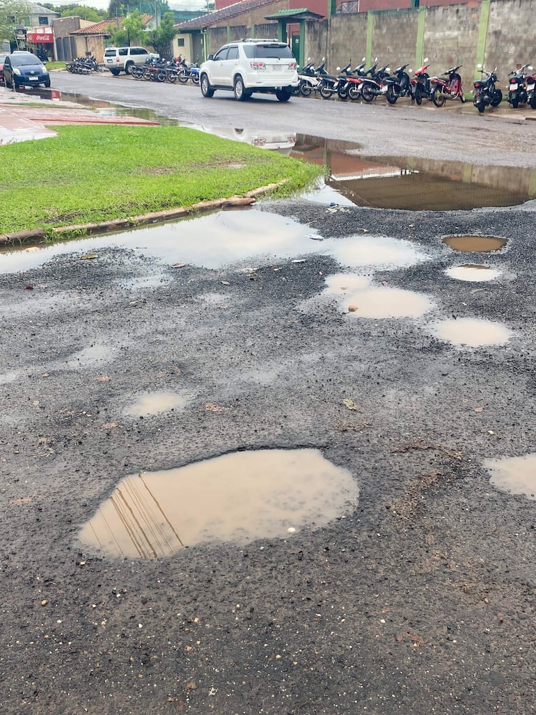 Calle con pavimento dañado y charcos de agua, mientras llueve. Motocicletas y vehículos estacionados a la derecha.