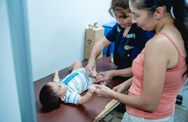 Mujer sostiene a un bebé en una mesa médica mientras otra profesional administra una inyección en un consultorio bien iluminado.