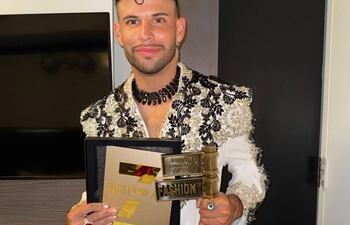 Simón Villalba, con chaqueta blanca y bordados, sostiene un premio y un documento en ambiente neutral.