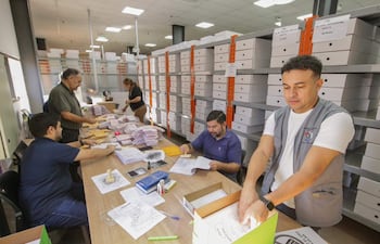 Hombre con chaleco gris coloca documentos en caja, rodeado de otros adultos revisando papeles en oficina ordenada.