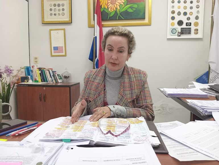 Irma Llano, mujer con abrigo a cuadros, sostiene documentos en su escritorio rodeada de libros y una planta.