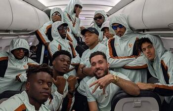 Jugadores del equipo francés de fútbol en avión, todos con chaquetas celestes, algunas personas usando gafas de sol y gorras, mostrando sonrisas.