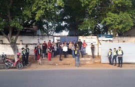 Grupo de personas frente a la escuela, algunas con ropa colorida, escuchando mientras una persona sostiene un cartel.