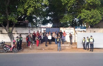 Grupo de personas frente a la escuela, algunas con ropa colorida, escuchando mientras una persona sostiene un cartel.