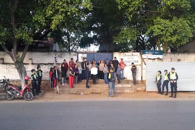 Grupo de personas frente a la escuela, algunas con ropa colorida, escuchando mientras una persona sostiene un cartel.
