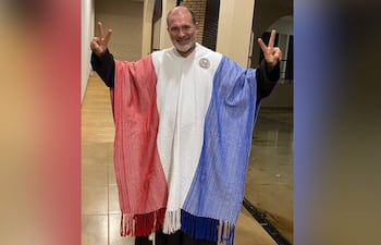 Sacerdote brasileño sonríe y muestra gestos de paz vistiendo un poncho de los colores de la bandera paraguaya en un espacio cerrado.