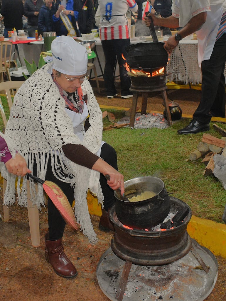 Mujer con poncho blanco y gafa blanca cocina en fogón, mientras un hombre la asiste. Ambiente festivo de cocina comunitaria.