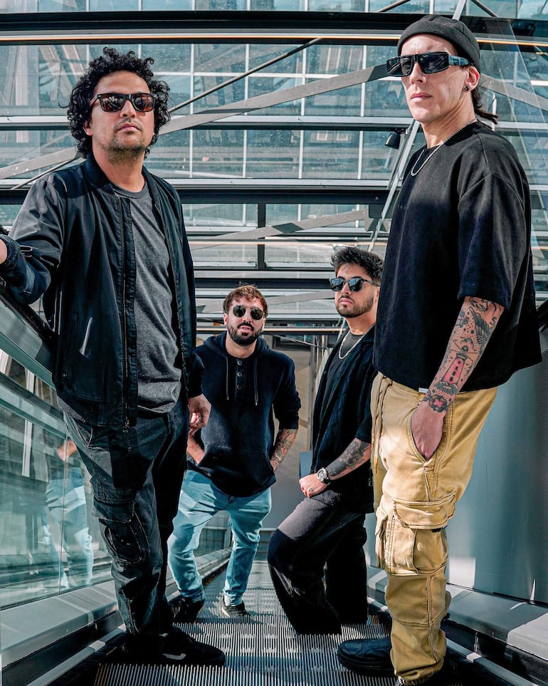 Cuatro hombres en escalera mecánica, con gafas de sol y ropa casual, posando en un ambiente moderno y relajado.