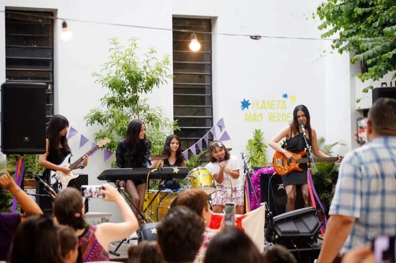 Grupo de cinco niñas en el escenario, una canta con micrófono y otras tocan guitarra, teclado y batería. Banderines morados decoran el entorno.