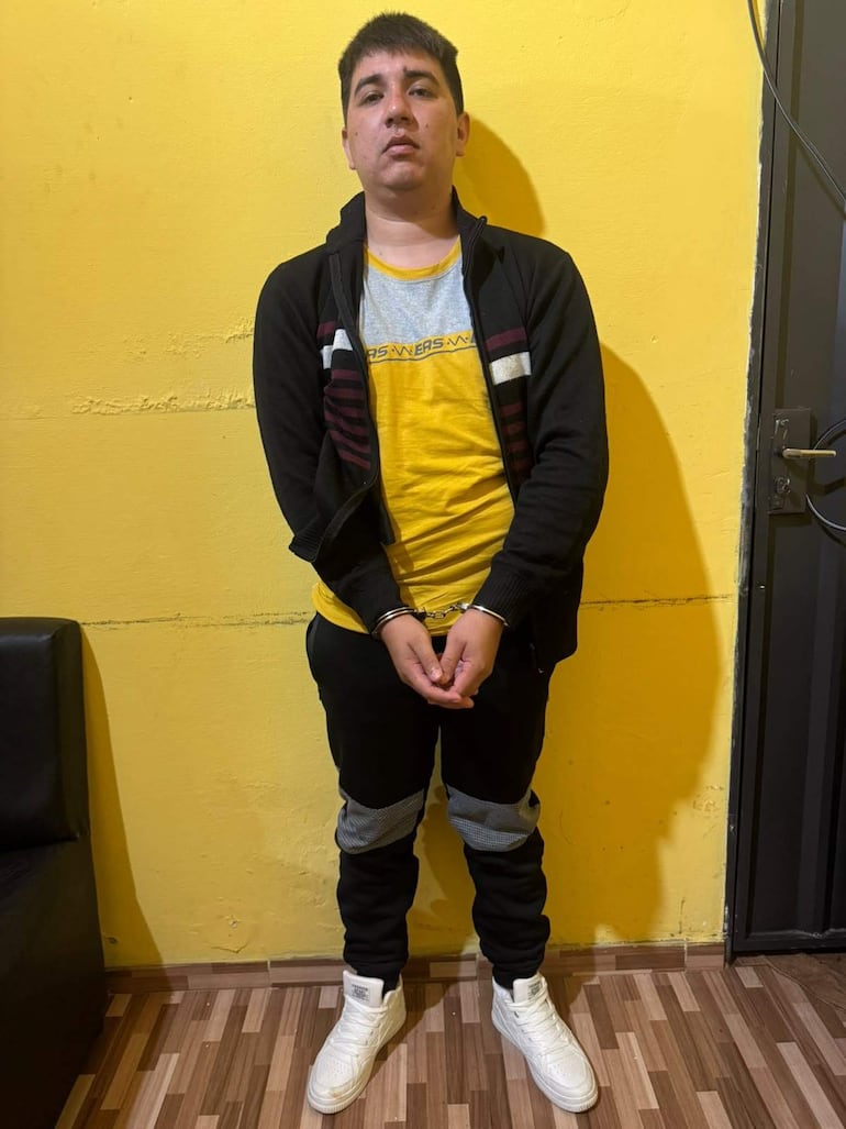 Hombre joven, de pie con manos esposadas, viste camiseta amarilla y gris, abrigo negro y pantalones oscuros en ambiente sencillo.
