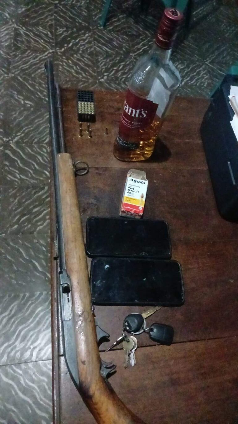 Mesa con escopeta, cartuchos, celulares y botella de whisky, en un interior con piso de mosaico.