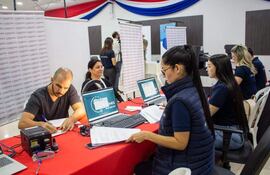 Cinco personas interactúan en un centro de votación; una mujer con chaleco azul observa una computadora mientras otros trabajan en laptops.