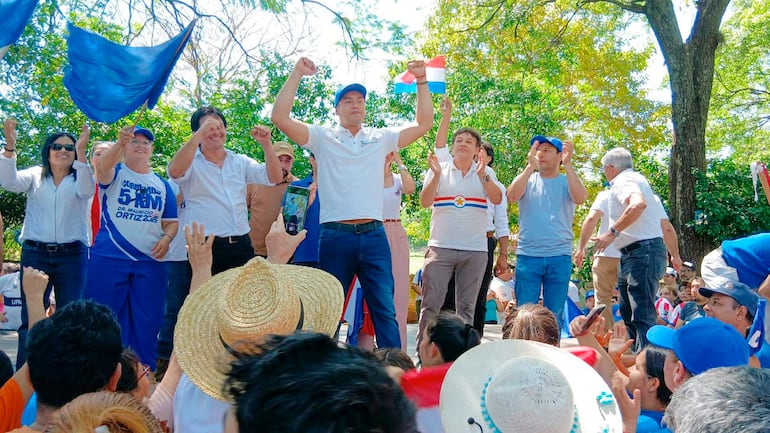 Miguel Prieto en escenario con brazos levantados, rodeado de personas aplaudiendo y agitando banderas de Paraguay, en un entorno natural.