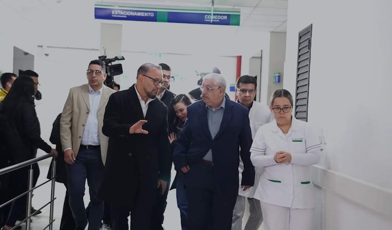 Isaías Ricardo Fretes en abrigo oscuro conversa con Derlis León en chaqueta clara, rodeados de personal médico en hospital.