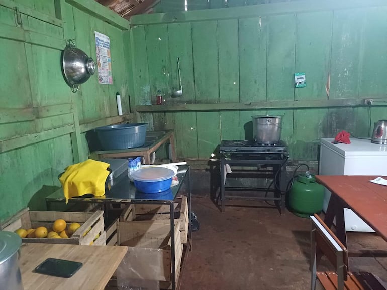 Ambiente de cocinas con paredes de madera verde, lavadero, mesa metálica y cajas de madera con naranjas en un lugar desatendido.