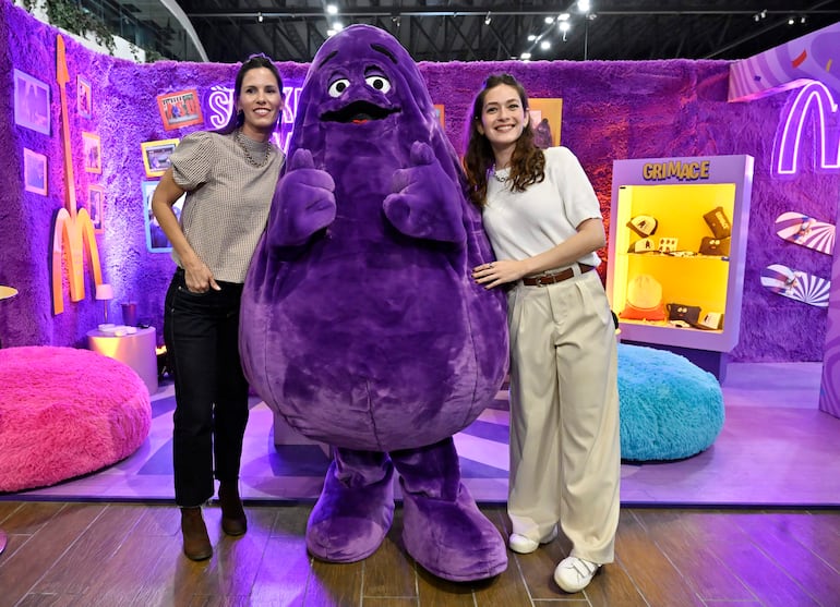 Mujeres sonrientes en blusas claras junto a Grimace, el personaje morado de McDonald's, en un ambiente festivo y colorido.