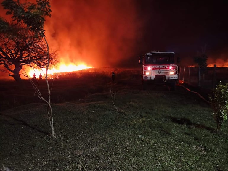 Dos personas cerca de intensas llamas y humo, con un camión de bomberos visible en el ambiente exterior anaranjado.