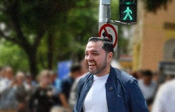Luis Bello sonríe, vistiendo camiseta clara y chaqueta oscura, cruzando una calle con otras personas en un día despejado.