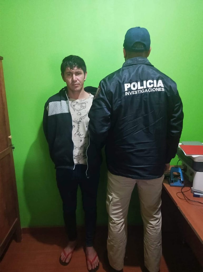 Hombre de contextura delgada con camiseta blanca y chaqueta oscura, detenido por policía en habitación con paredes verdes.