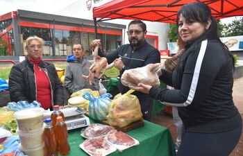 Mujer en suéter rojo observa a un hombre con pollo crudo y una mujer con carne envuelta, en feria bajo toldo rojo.
