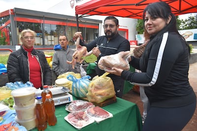 Mujer en suéter rojo observa a un hombre con pollo crudo y una mujer con carne envuelta, en feria bajo toldo rojo.