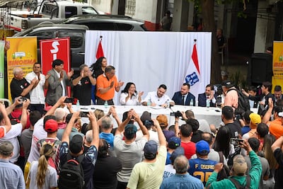 Luis Bello firmando documentos en un escenario, rodeado de trabajadores que aplauden. Ambiente festivo con banderas al fondo.