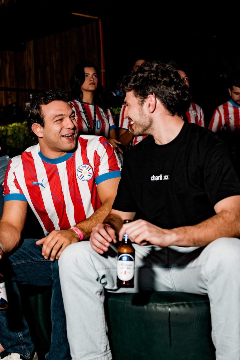 Dos hombres riendo, uno en camiseta blanca con franjas rojas y otro en camiseta negra, sosteniendo botellas de cerveza en un entorno festivo.