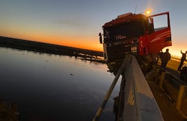 Camión rojo en el puente Nanawa, baranda a un lado, con el atardecer de fondo y un río fluyendo debajo.