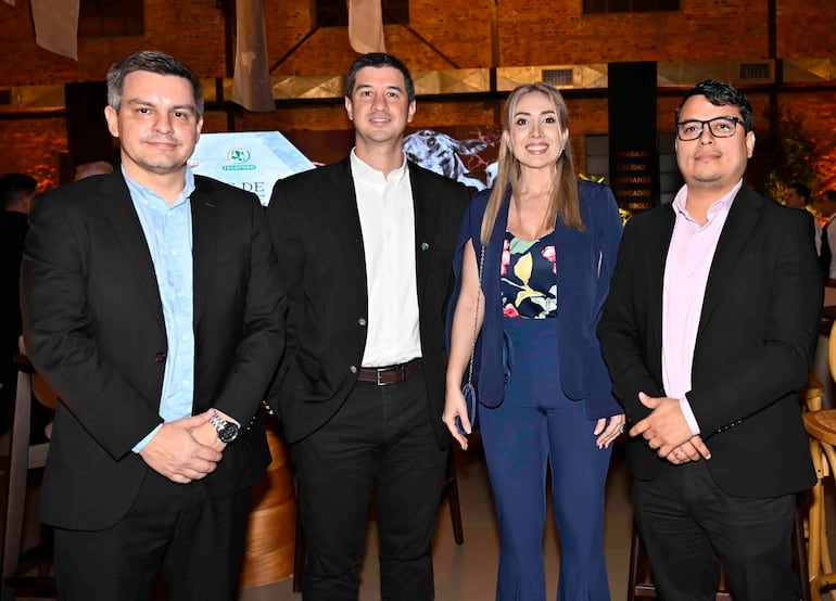 Cuatro personas sonrientes: un hombre con saco negro, una mujer elegante en azul, y dos hombres con sacos oscuros, en un ambiente moderno.