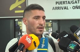 Hombre de cabello corto en abrigo gris y camiseta clara, rodeado de micrófonos en un entorno de aeropuerto.