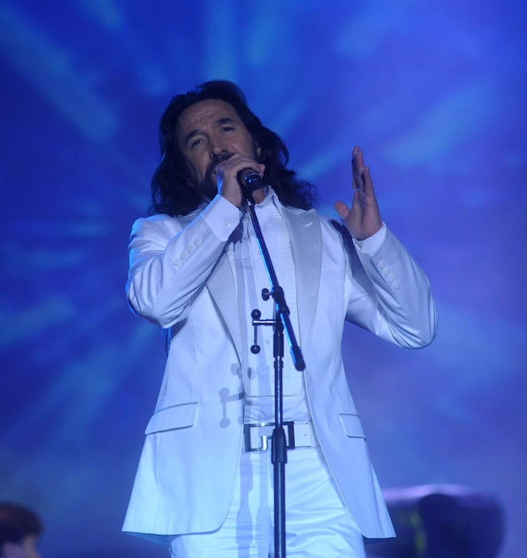 Marco Antonio Solís en traje blanco, cantando solo en el escenario iluminado, con expresión tranquila y micrófono en mano.