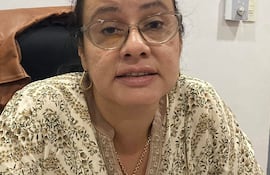 Mujer con top floral y gafas, sentada y en conversación, fondo de pared blanca y asiento oscuro.