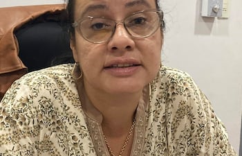 Mujer con top floral y gafas, sentada y en conversación, fondo de pared blanca y asiento oscuro.