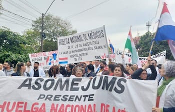 Grupo de médicos con camisetas blancas sostiene un gran cartel de ASOMEJUMS, mientras otros muestran pancartas de reclamos en un ambiente urbano.