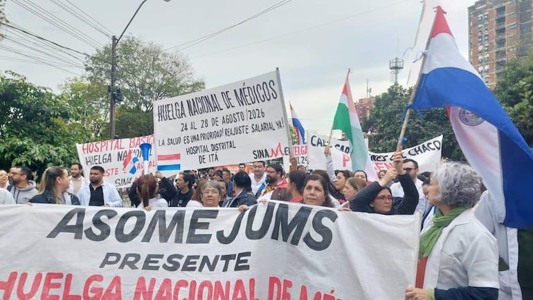 Grupo de médicos con camisetas blancas sostiene un gran cartel de ASOMEJUMS, mientras otros muestran pancartas de reclamos en un ambiente urbano.