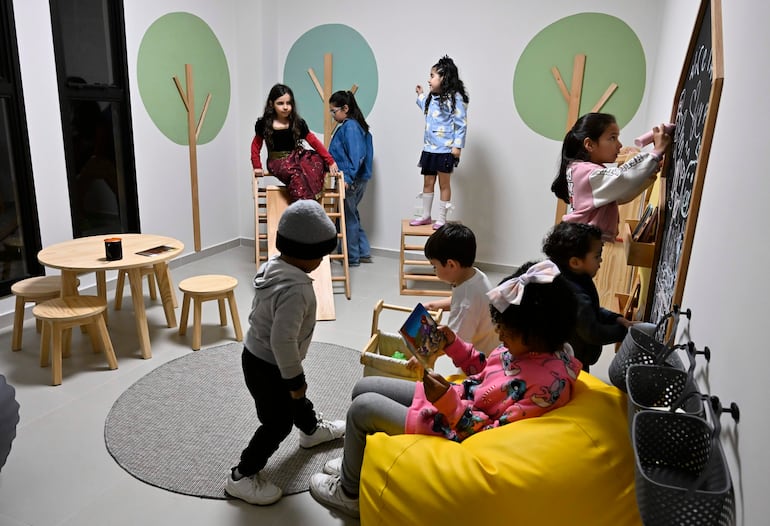 Niños interactuando en un salón con paredes blancas, juguetes y decoración colorida; una niña en vestido negro y chal rojo se destaca.