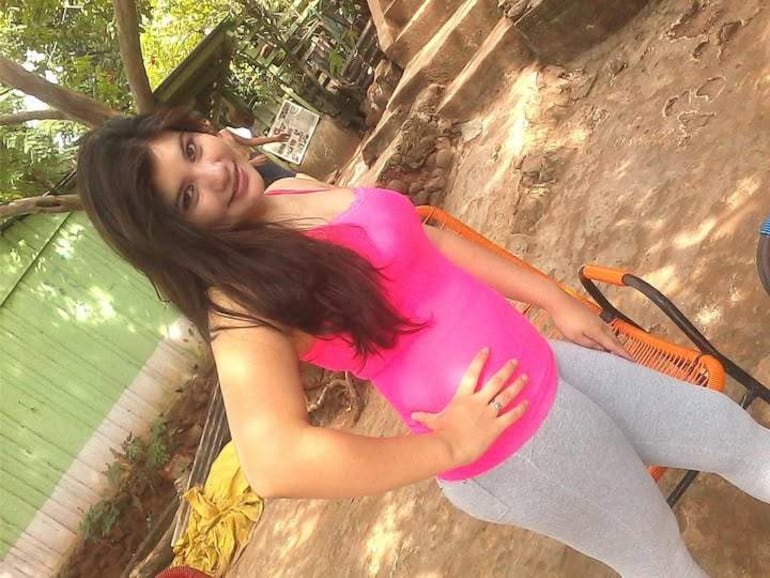 Mujer de pie con blusa rosa y leggings grises, sonriendo hacia la cámara en un patio con vegetación.