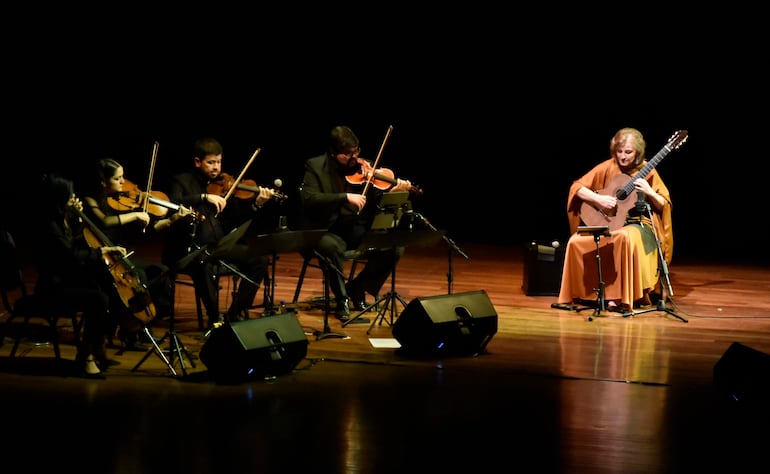 Berta Rojas, con cabello rubio y prenda naranja, toca la guitarra en un escenario con músicos de vestimenta oscura y luces tenues.