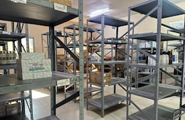 Almacén de medicamentos con estanterías grises parcialmente llenas de cajas y frascos, sin personas visibles en la imagen.