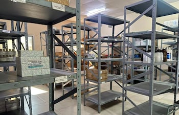 Almacén de medicamentos con estanterías grises parcialmente llenas de cajas y frascos, sin personas visibles en la imagen.