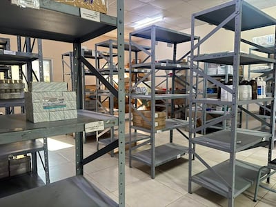 Almacén de medicamentos con estanterías grises parcialmente llenas de cajas y frascos, sin personas visibles en la imagen.