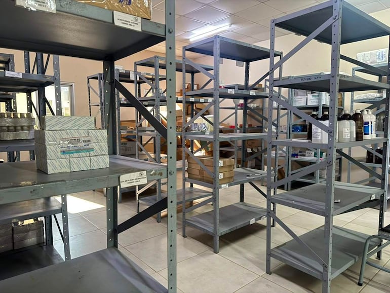 Almacén de medicamentos con estanterías grises parcialmente llenas de cajas y frascos, sin personas visibles en la imagen.
