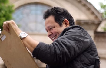 Carlos Sosa, hombre de cabello corto y chaqueta negra, sonríe mientras dibuja en un panel de madera en un ambiente urbano.