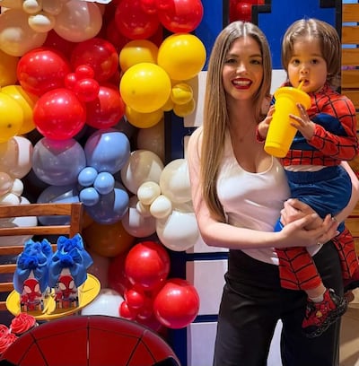 Mujer con camiseta blanca sonríe mientras sostiene a un niño en traje de Spider-Man, rodeados de globos coloridos.
